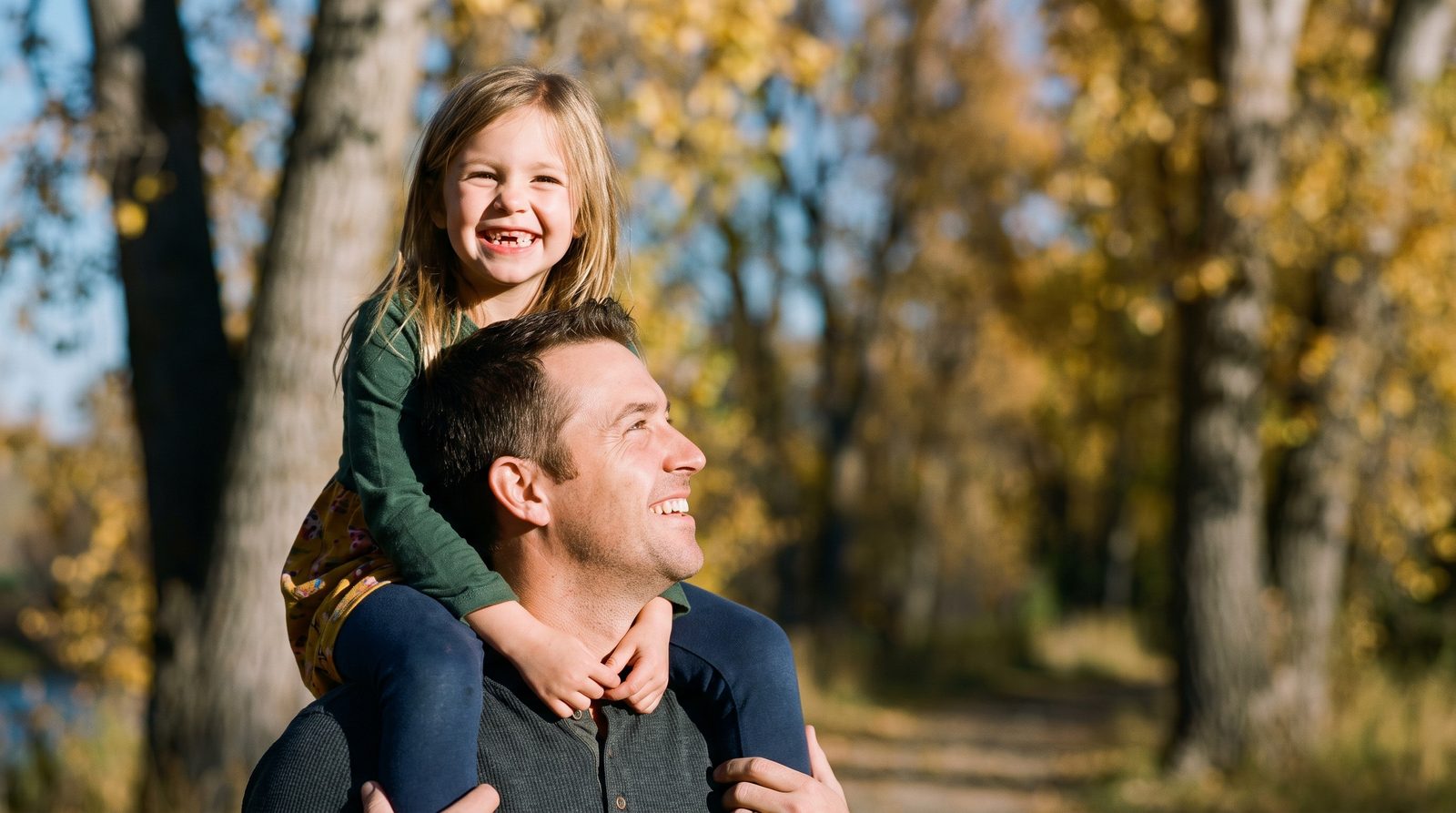 Papá cargando en hombros a su hija, a la que le falta un diente, en un sendero junto al río en otoño